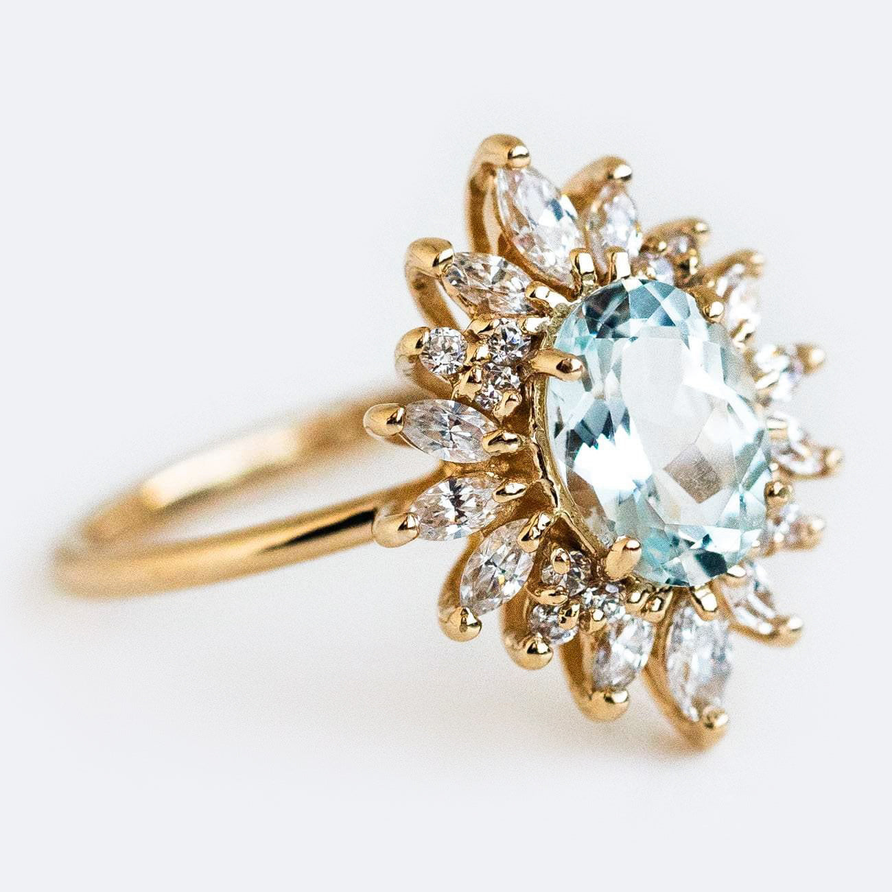 The Lana Ring with Aquamarine & CZ is crafted in 14k solid gold, showcasing an oval-cut pale blue aquamarine center stone surrounded by marquise and round clear cubic zirconia in a floral sunburst halo design.