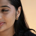 Close-up of a woman with long black hair and smooth skin, smiling softly. Her left ear is adorned with a shiny Cosmic Tapered Huggie Earring in 14k yellow gold plating. The background is warm beige.