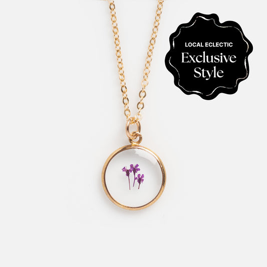 The Tiny Birth Flower Portrait Necklace is a gold necklace with a round pendant featuring pressed purple flowers. A black label in the top right corner reads Local Eclectic Exclusive Style.