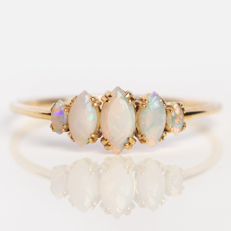 The Solid Gold Essential Opal Marquise Cluster Ring (sample size 7) features five marquise and oval opals in a row, their iridescent pastel hues showcased on a light background with a subtle reflection beneath.