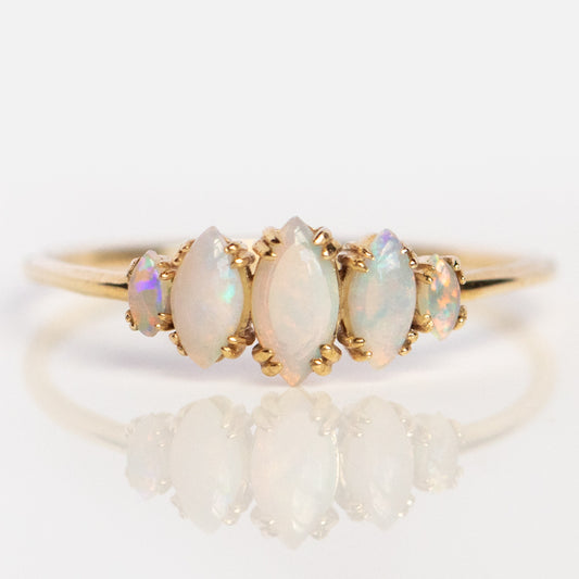 The Solid Gold Essential Opal Marquise Cluster Ring (sample size 7) features five marquise and oval opals in a row, their iridescent pastel hues showcased on a light background with a subtle reflection beneath.