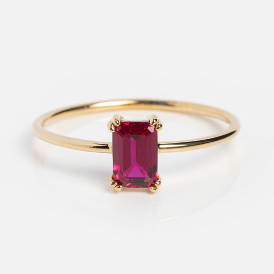 The Solid Gold Essential Baguette Birthstone Ruby Ring Sample features a thin 9k yellow gold band (1mm) with a rectangular, emerald-cut deep pink ruby set in simple prongs, shown on a white background.
