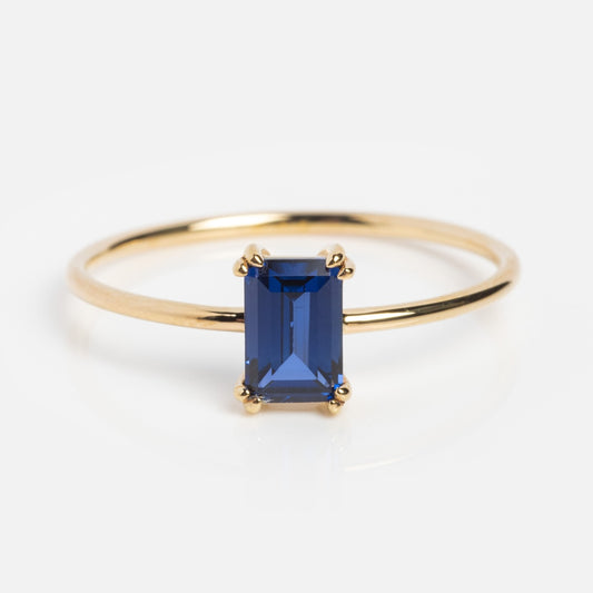 The Solid Gold Essential Baguette Birthstone Ring features a slim gold band with a rectangular, emerald-cut deep blue gemstone in a four-prong setting, shown on a white background.