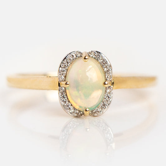 The Solid Gold October Capsule Oval Opal Ring Size 8 Sample features an oval opal center with a halo of round white diamonds, crafted in 9k yellow gold and displayed on a reflective white surface.