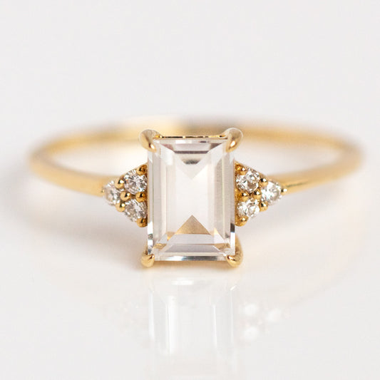 The Solid Gold Essential White Topaz Ring Sample features a rectangular emerald-cut white topaz center stone, accented by three round white diamonds on each side, and is crafted in yellow gold, shown on a white surface.