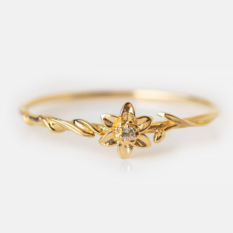The Solid Gold Birth Flower Ring features a delicate flower design with a central round diamond, set on a thin, twisted gold band, shown against a plain white background.
