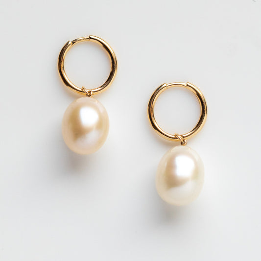 The Solid Gold Elegant Pearl Charm Huggies feature large, oval freshwater pearls dangling from solid gold hoops, shown against a plain white background.