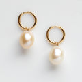The Solid Gold Elegant Pearl Charm Huggies feature large, oval freshwater pearls dangling from solid gold hoops, shown against a plain white background.