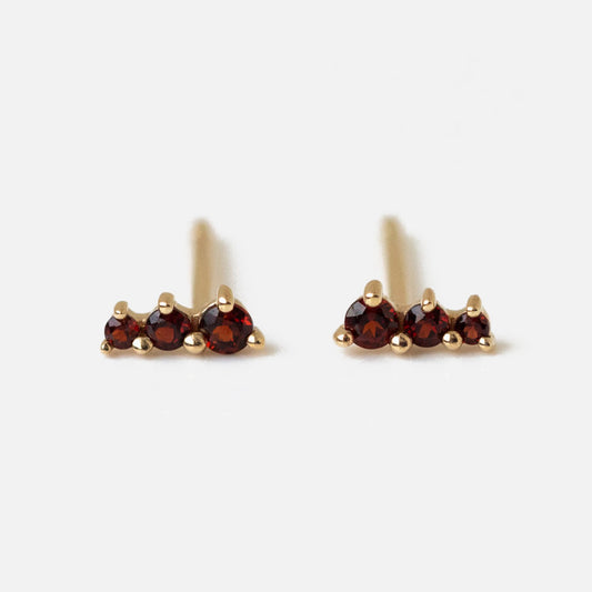 A pair of Solid Gold Dainty Birthstone Bar Studs, each adorned with three small round garnet gemstones arranged in a row, displayed against a plain white background.