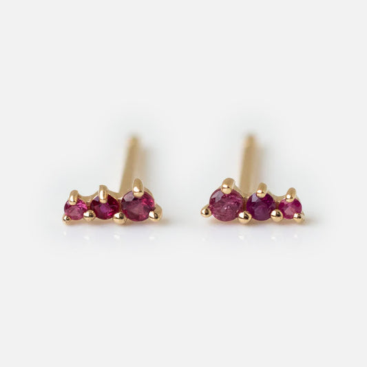 A pair of Solid Gold Dainty Birthstone Bar Studs, each featuring three small round red gemstones in a row, is displayed on a white background.