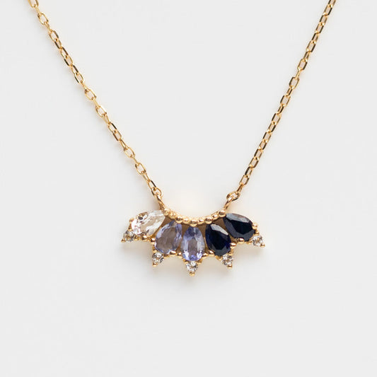 The Solid Gold Ombre Birthstone Necklace September Sample is a 9k yellow gold chain with a fan-shaped pendant featuring five teardrop gemstones in blue and clear tones, including white topaz, accented with small stones at the base.