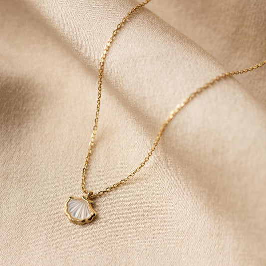 The Solid Gold Mother of Pearl Shell Necklace Sample is a delicate 9k yellow gold chain with a scallop shell pendant, featuring a glossy mother of pearl center outlined in gold, shown resting on soft beige satin.