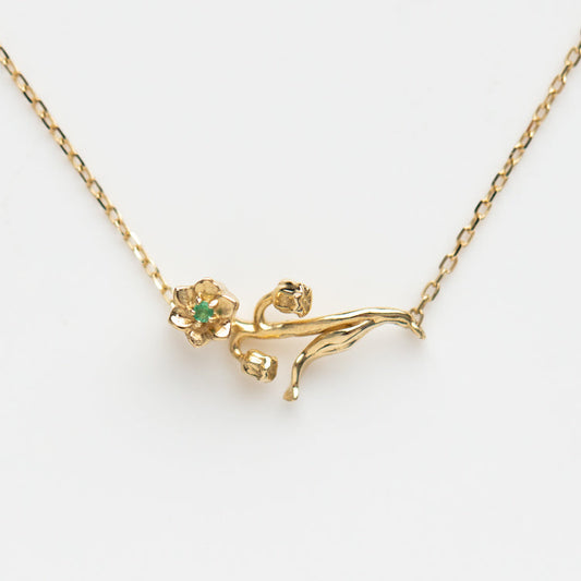 The Solid Gold Birth Flower Necklace May Sample features a delicate 9k yellow gold pendant shaped like a reclining figure holding a flower with a natural emerald center, displayed against a plain white background.