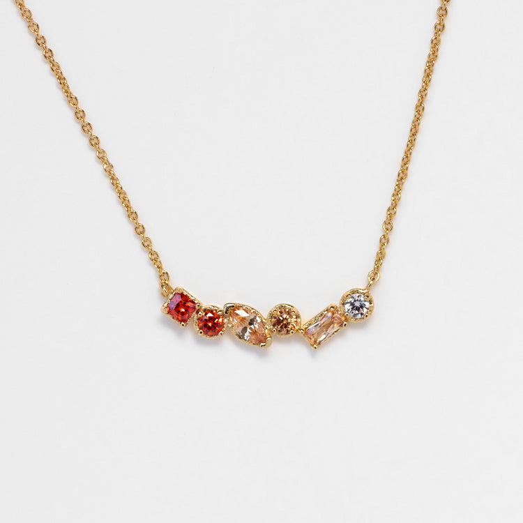The Shades of You Ombre Birthstone Necklace is a delicate gold necklace featuring five uniquely shaped and colored gemstones in a row, set on a simple gold setting against a white background.