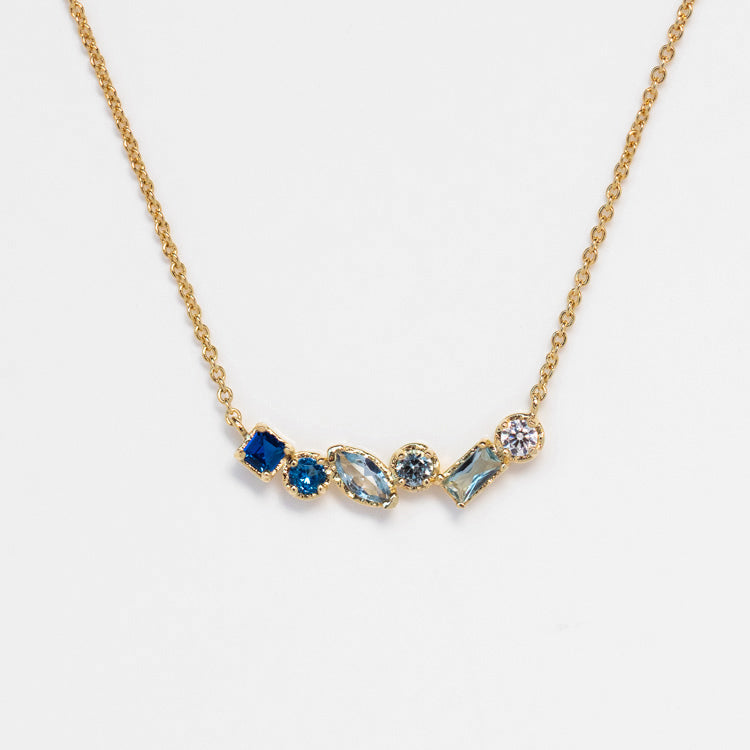 The Shades of You Ombre Birthstone Necklace features five uniquely cut blue and clear gemstones set in a row on a gold chain, creating an elegant ombre effect, shown against a white background.