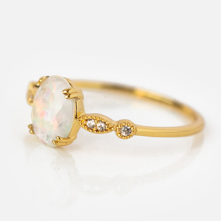 The Opal Fire Ring showcases a delicate gold band with a round, faceted simulated opal center and two small diamonds on each side, displayed against a plain white background.
