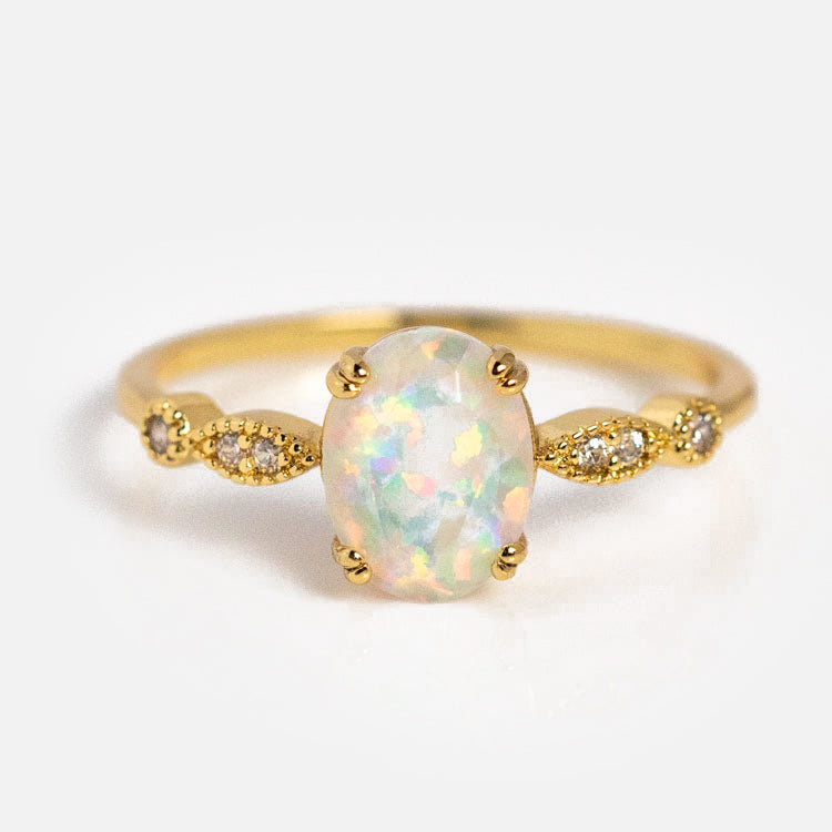 The Opal Fire Ring features a central simulated opal gemstone, accented by small round diamonds set in decorative gold details on each side, displayed against a plain white background.