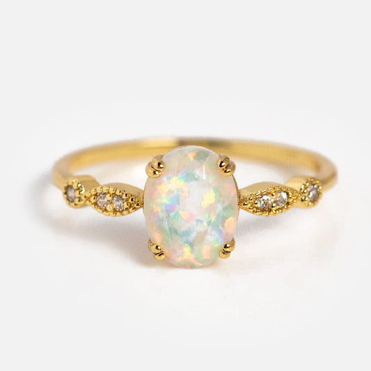 The Opal Fire Ring features a central simulated opal gemstone, accented by small round diamonds set in decorative gold details on each side, displayed against a plain white background.