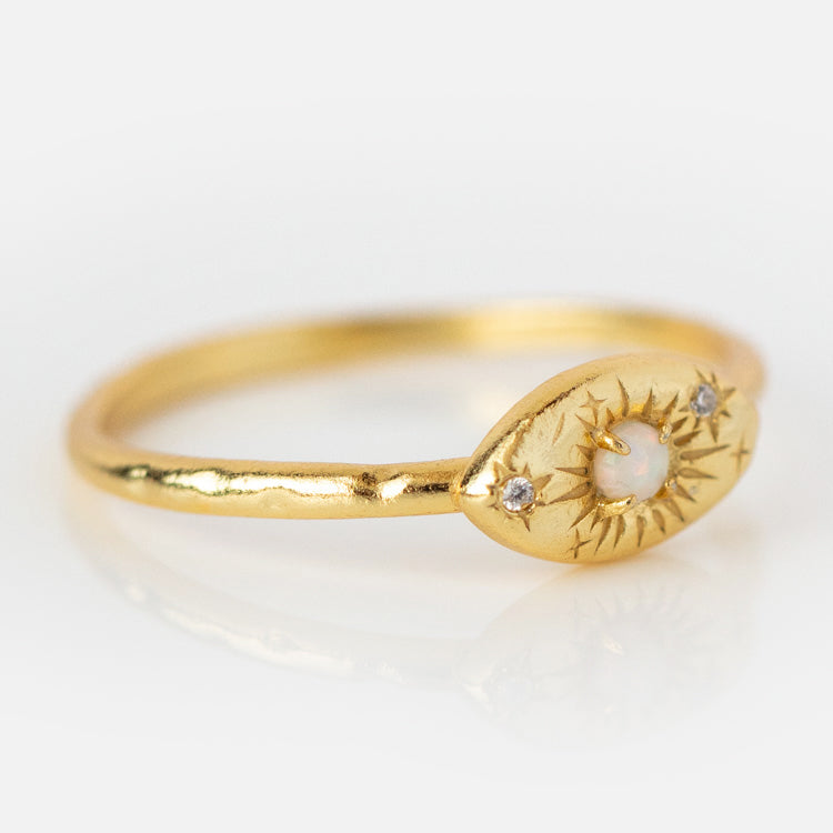 The Opal Amulet Ring is a delicate gold-plated piece with a thin band, an oval face engraved with a starburst pattern, featuring a central opal and two small diamonds on the sides.