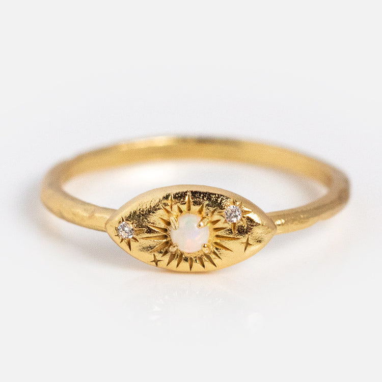 The Opal Amulet Ring is a delicate gold plated band with an eye-shaped design, central opal stone, two accent diamonds, and engraved starburst details, displayed on a plain white background.
