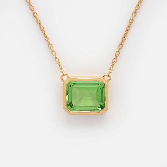 The Around the Sun Birthstone Necklace is a yellow gold plated chain with a square-cut vivid green gemstone pendant in a gold bezel, shown on a white background. Free gift with orders over $200.