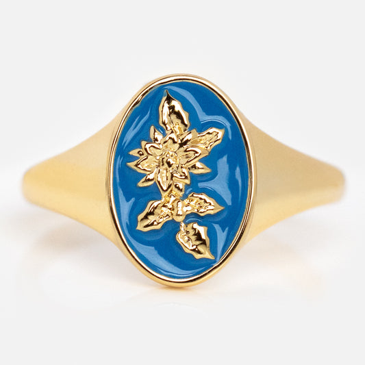 The Enamel Birth Flower Signet Ring features an oval blue enamel face with a raised gold floral design at the center and a polished gold-plated band for a modern, elegant look.