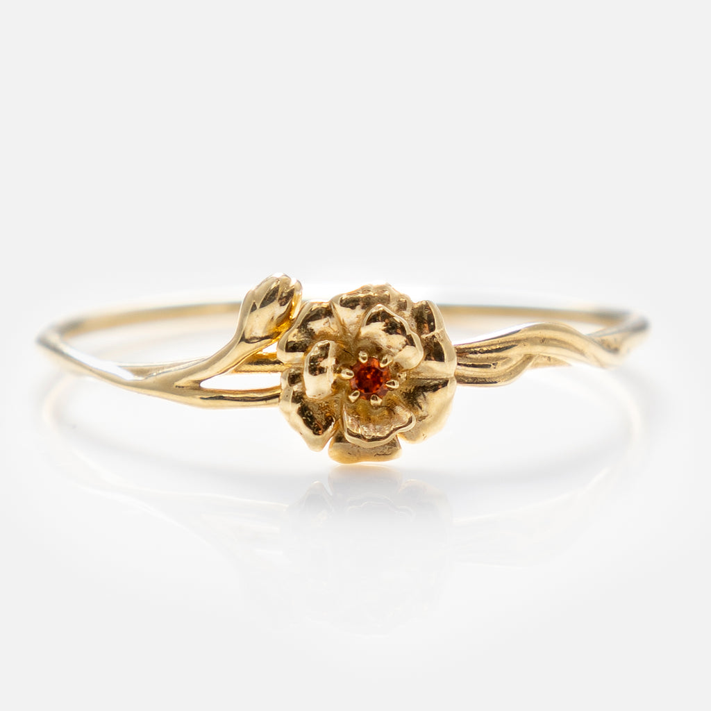 The Solid Gold Birth Flower Ring features a detailed flower design at the center with a small red gemstone, set against a plain white background.