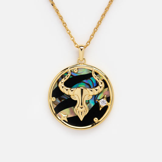 The Zodiac Pendant Necklace features a 14k gold plated chain with a round pendant showcasing a gold bull’s head over an iridescent background, detailed with a star, gold accents, and a clear crystal—ideal as a zodiac accessory.