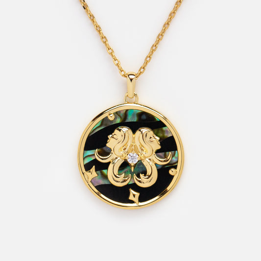 The Zodiac Pendant Necklace features a 14k gold plated round pendant with an ornate gold Gemini sign, a central diamond, and an abalone shell background with iridescent green and blue tones.