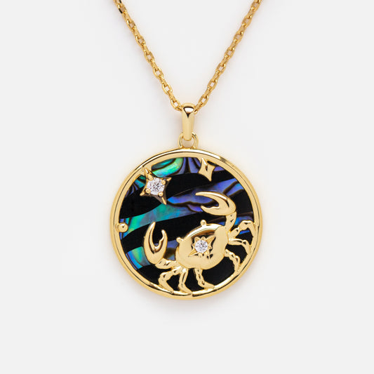 The Zodiac Pendant Necklace is a 14k gold plated piece with a round crab zodiac pendant, two sparkling stones, and an iridescent blue-green inlay.