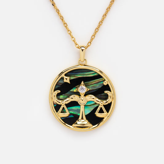 The Zodiac Pendant Necklace features a 14k gold-plated round pendant with a scales of justice and star design, set against an iridescent abalone shell background.