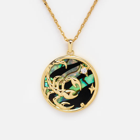 The Zodiac Pendant Necklace features a 14k gold plated round pendant with an intricate gold Scorpio symbol on iridescent abalone shell, accented by a sparkling gemstone for a celestial touch.