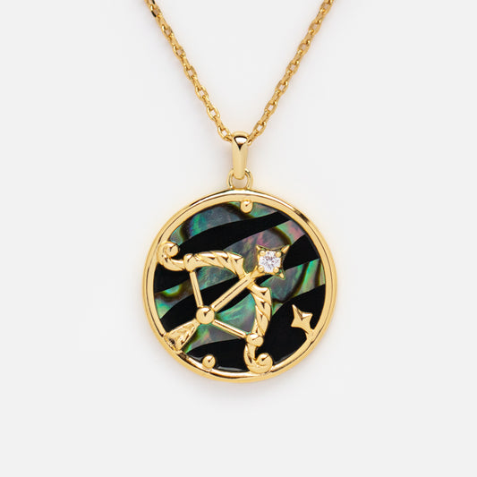 The Zodiac Pendant Necklace is a 14k gold plated piece with a round constellation pendant, bow and arrow design, iridescent abalone shell inlay, black accents, and a sparkling clear gemstone.