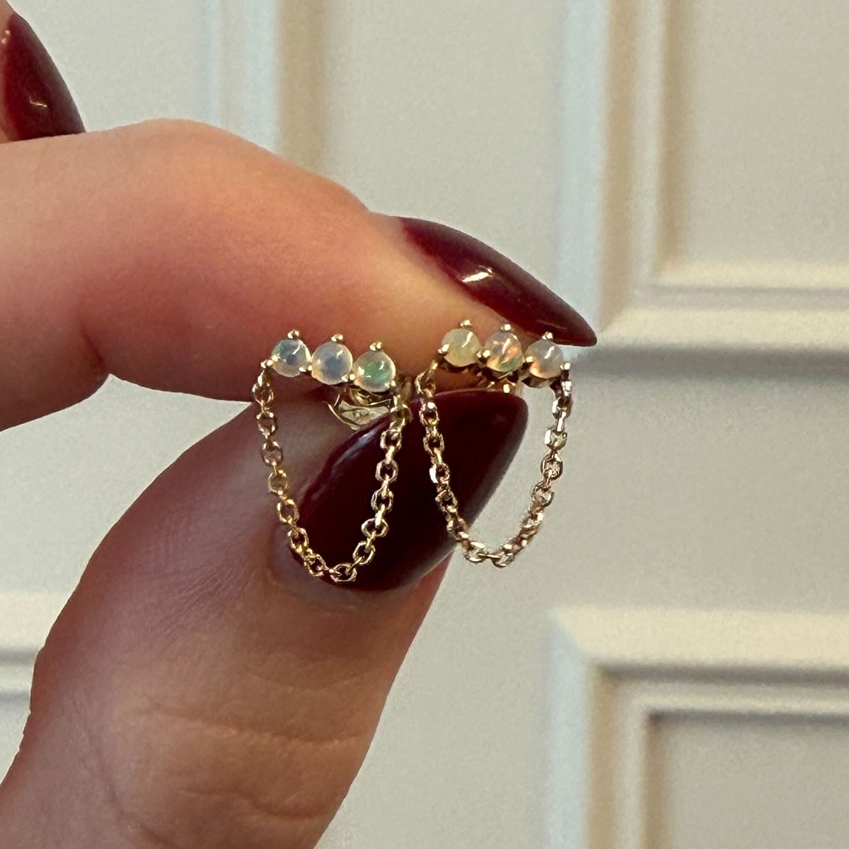 A hand with dark red nails holds the Solid Gold Opal Trio Chain Dangle Studs, featuring a delicate gold chain and three Ethiopian opals in a row, with a softly blurred white-paneled background highlighting their elegance.