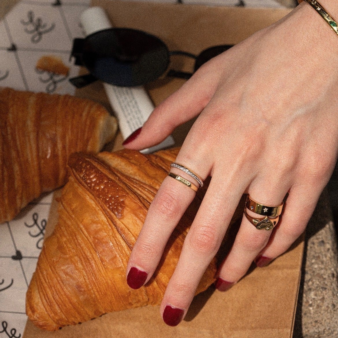 A hand with red-painted nails, wearing the Tiny Cunt Signet Ring, 14k gold-plated bracelets, and silver rings, rests on a croissant. Also visible are another croissant, sunglasses, and a paper bag.
