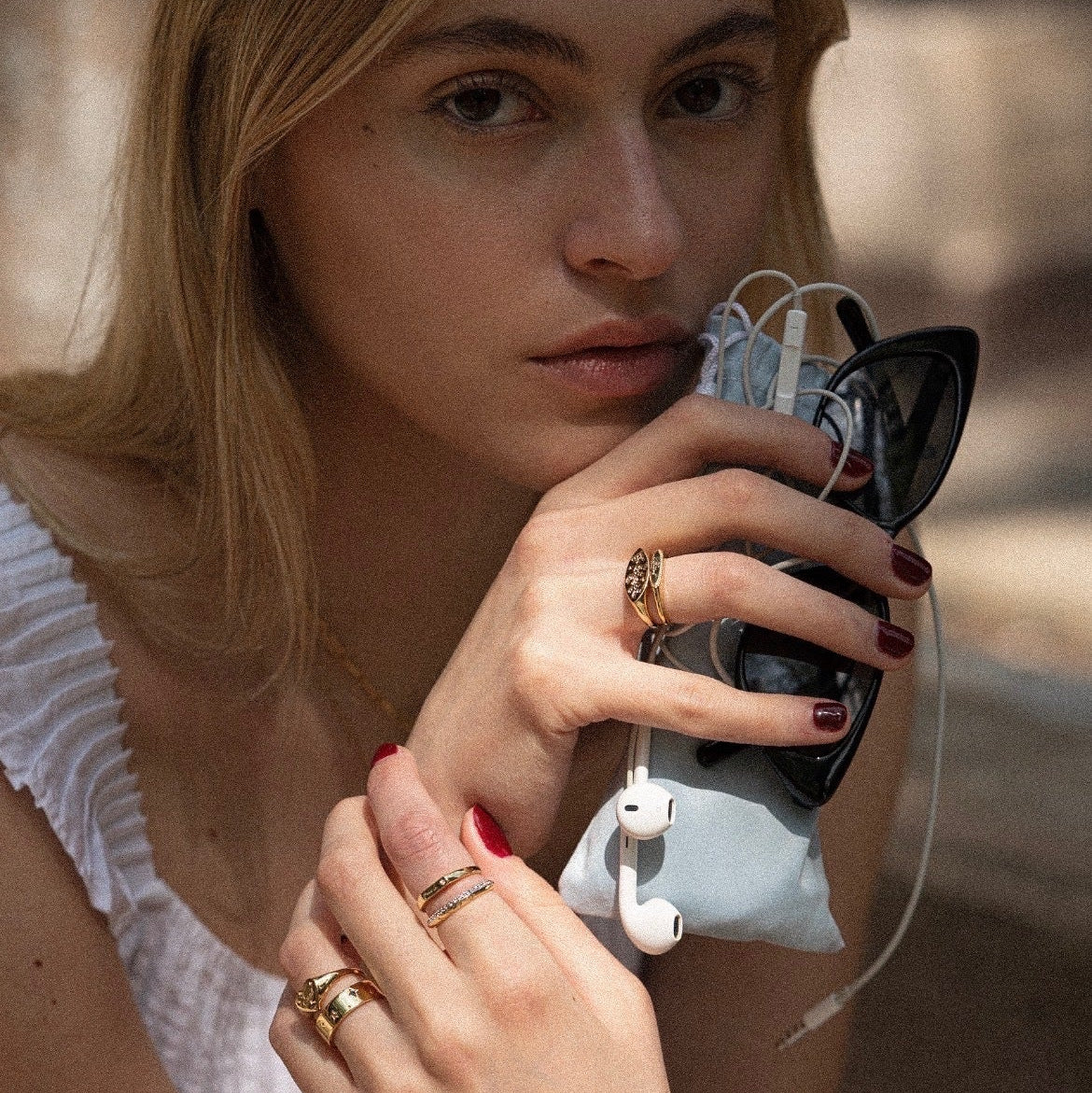 A woman with long blonde hair and a white top holds sunglasses, wired earphones, and a pouch, showing off her red nails and the Tiny Cunt Signet Ring in natural light while looking at the camera.