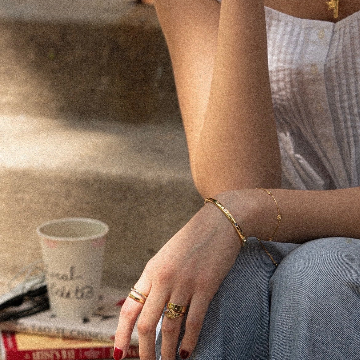 Wearing the Tiny Cunt Signet Ring and 14k gold plated bracelets, a person in a white sleeveless top sits on steps, arm on knee. A paper coffee cup and two books are beside them as sunlight creates soft shadows.