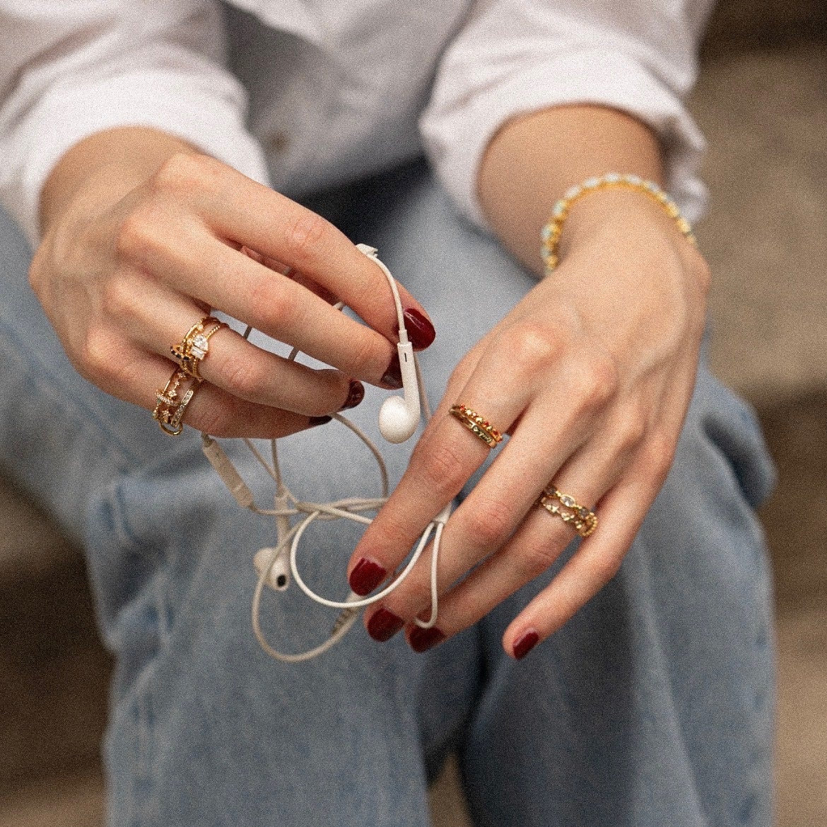 A person with red nail polish wears the Starlet Infinity Stacking Ring and a beaded bracelet while holding white earphones, dressed in a white shirt and blue jeans.