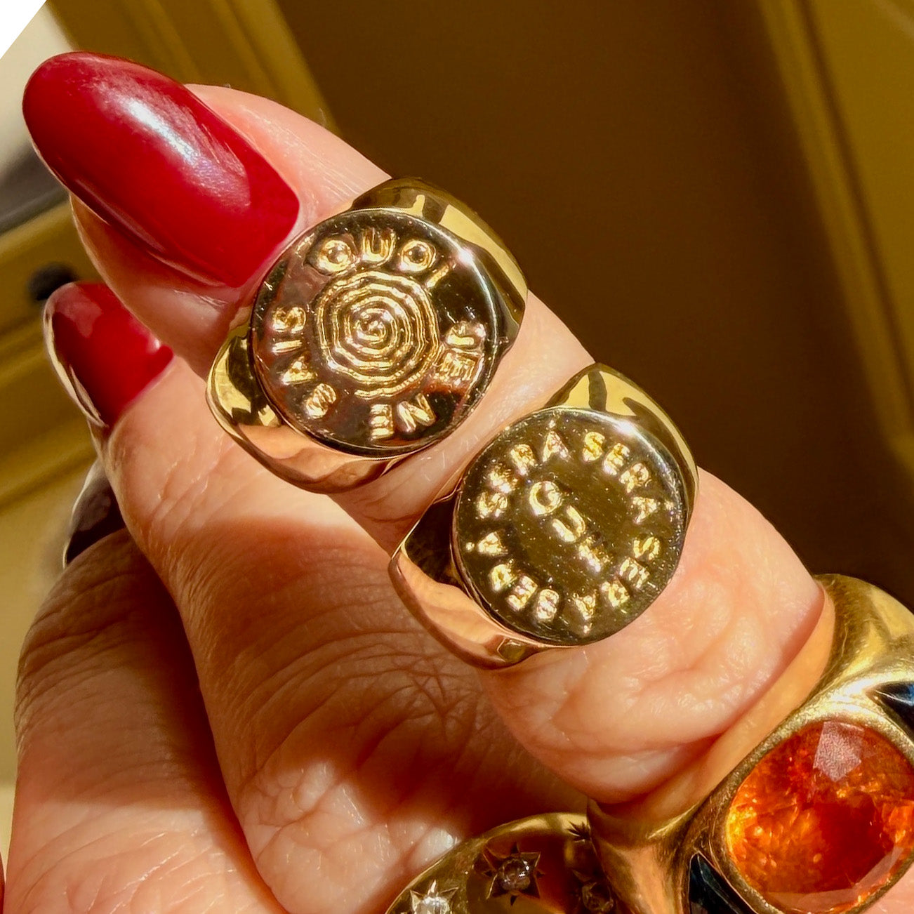 A hand with red-painted nails displays two gold plated rings—one a spiral signet engraved SANG RUDO JE ENE and another reading QUE SERA SERA. The hand is also adorned with the I Like It Here Club x Local Eclectic Je Ne Sais Quoi Pinky Ring.