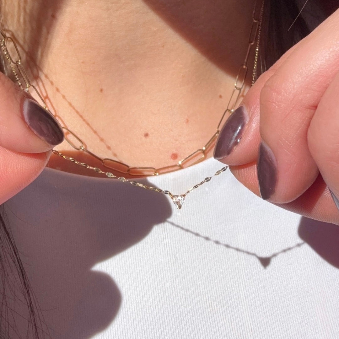 A person with painted nails holds the Solid Gold White Topaz You Have My Heart Necklace, featuring a delicate pendant. They wear a white top and layered gold chains, as sunlight casts soft shadows on their skin.