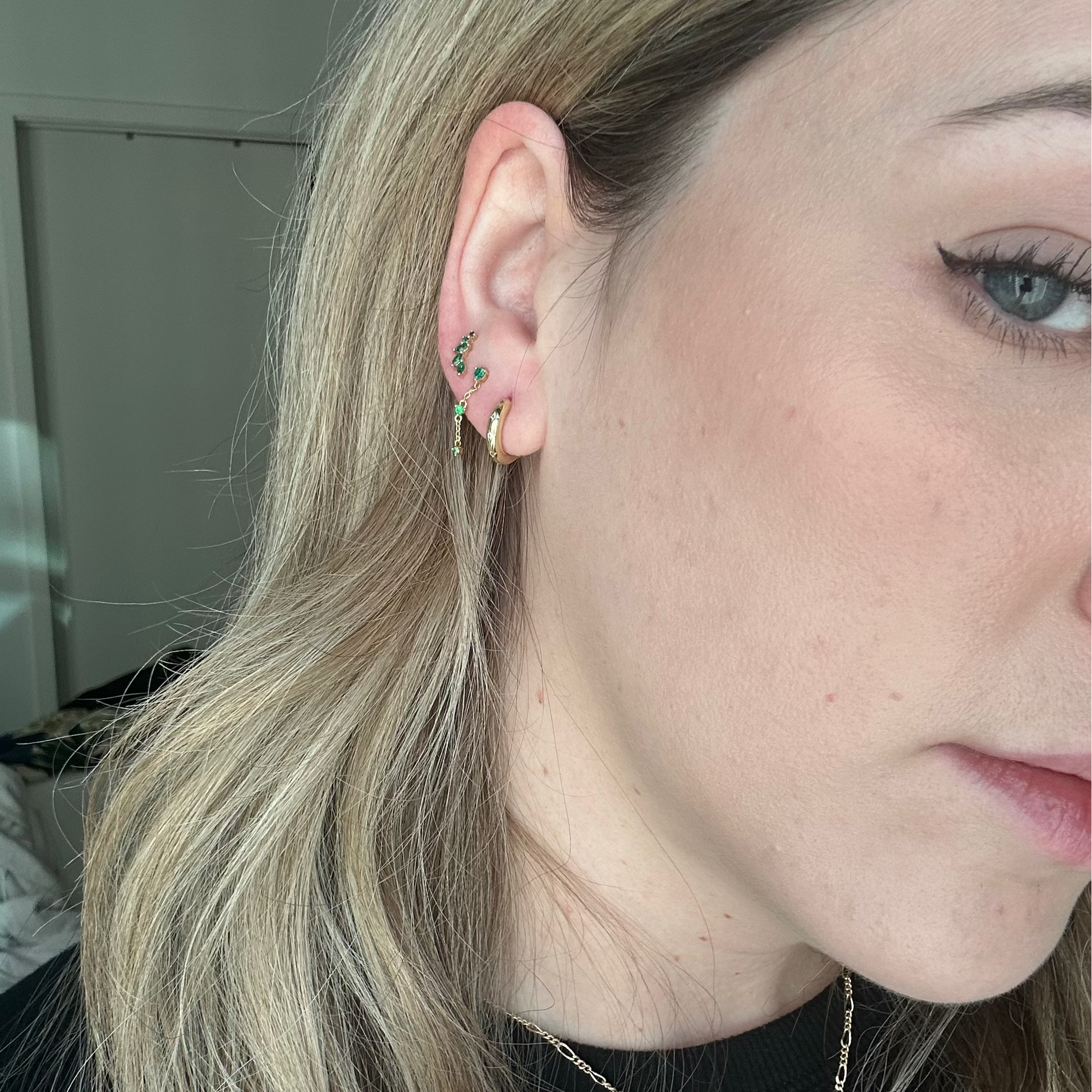 A close-up of a woman with light skin and blonde hair shows her ear adorned with the Local Eclectic exclusive Classic Birthstone Earring Set, featuring four gold and green gemstone earrings, against an indoor background with white walls.