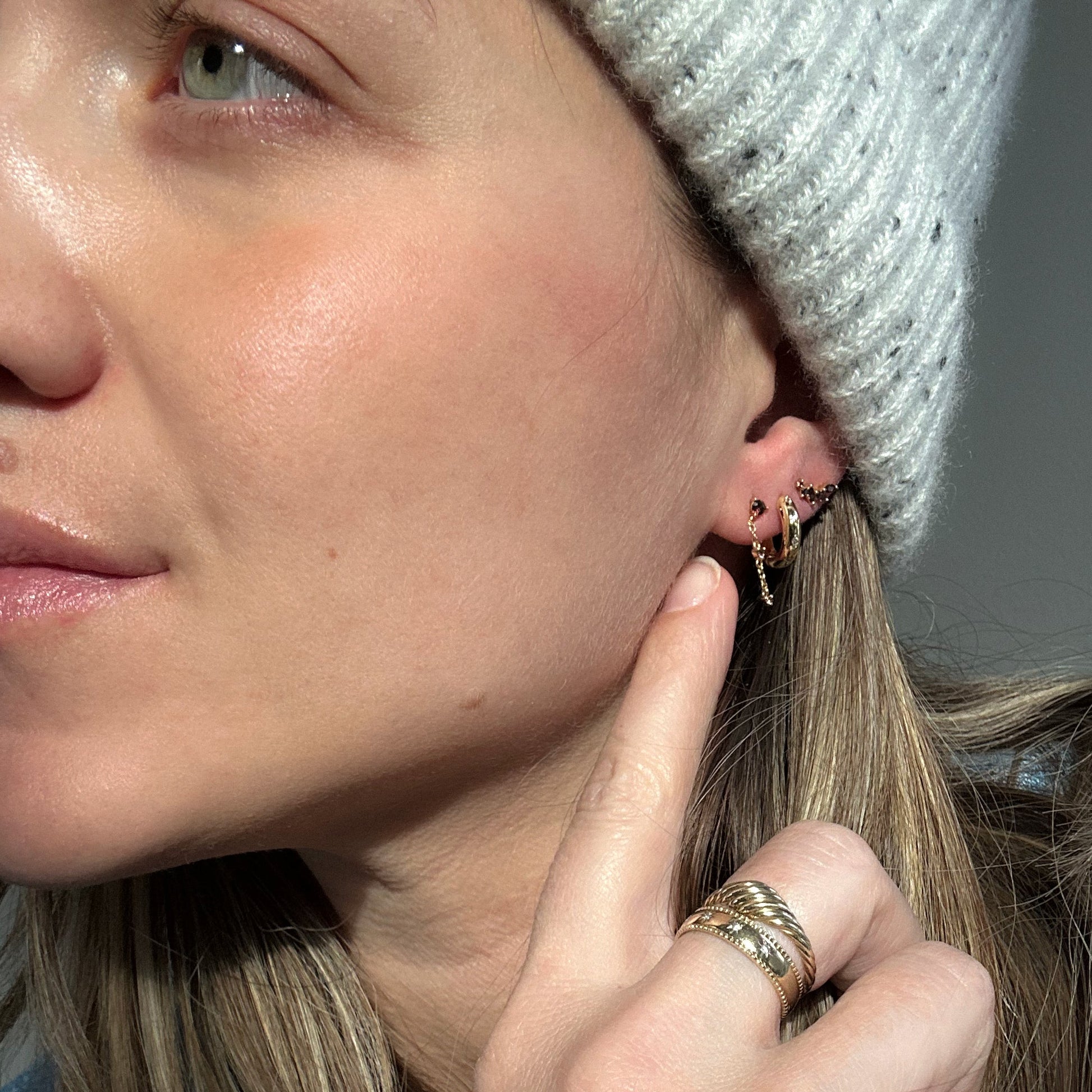 Close-up of a woman wearing a white knit hat, gold rings, and the Classic Birthstone Earring Set—14k gold plated accents catch the sunlight as she touches her ear, highlighting her personalized ear stack.