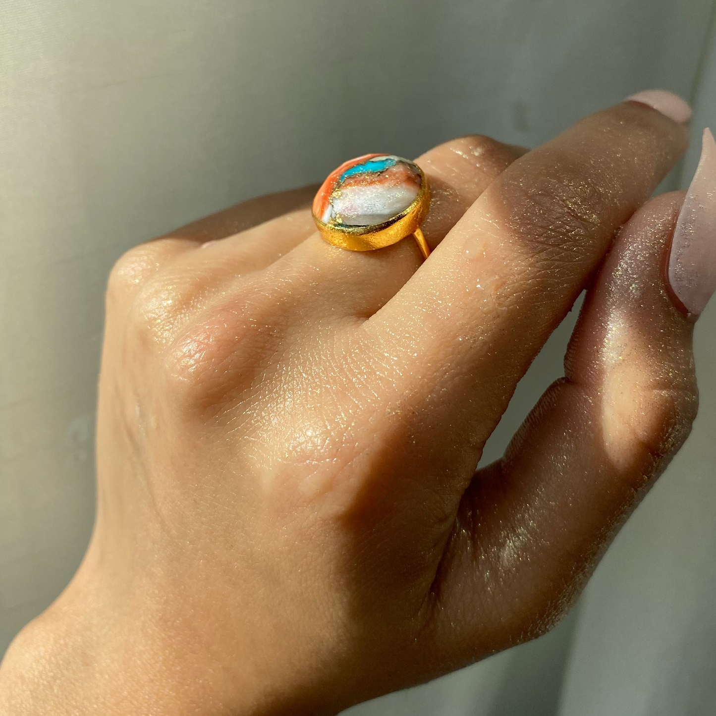 A hand with light brown skin wears the Carrie Elizabeth Statement Oyster Turquoise Ring, featuring a colorful oval stone, set against a softly lit neutral background.