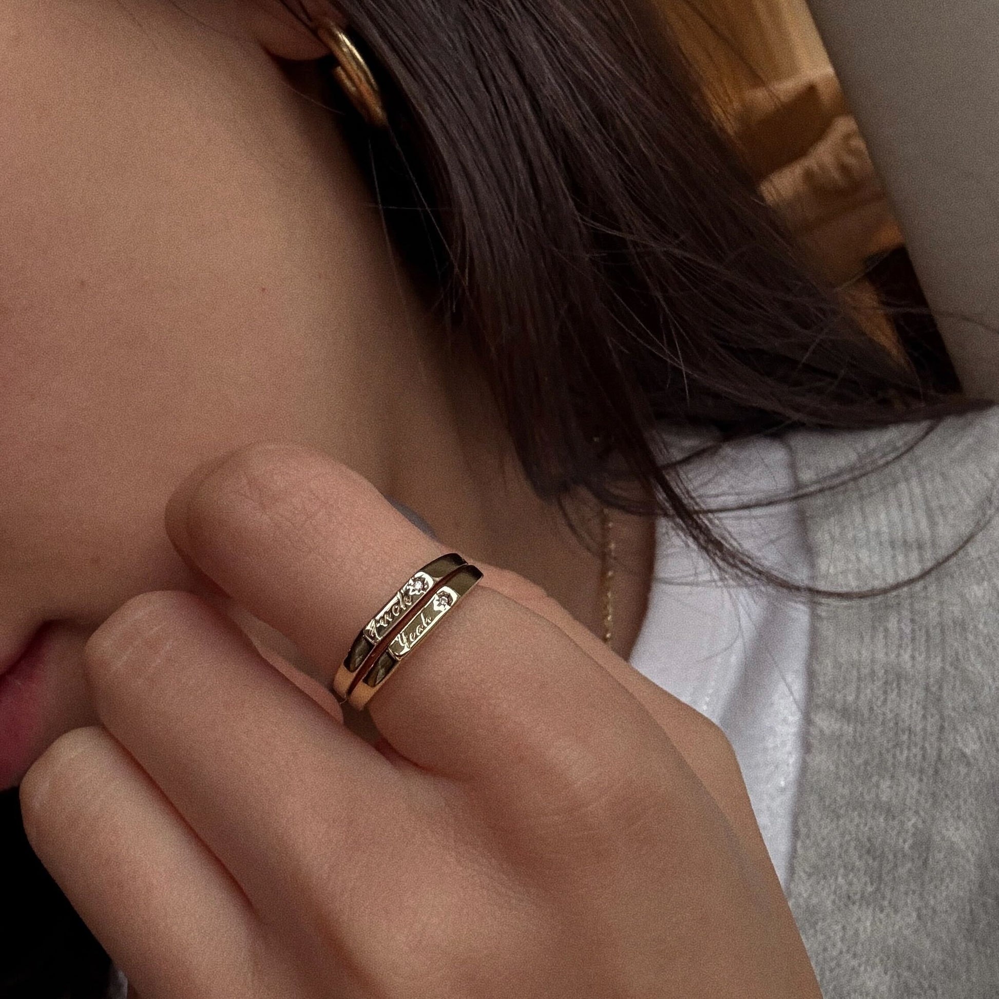 A close-up shows a hand with the Tiny Conversation Stacking Ring—14k yellow gold plated with small diamonds—on the ring finger, paired with a gold hoop earring. The person wears a grey sweater over a white top.