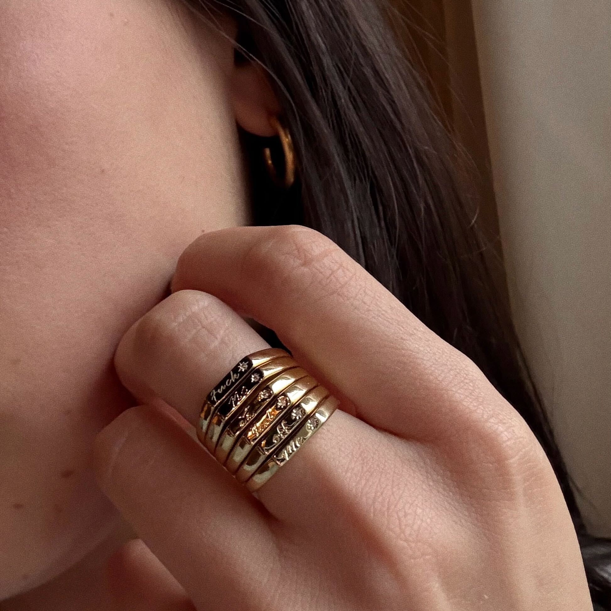 A close-up of a hand wearing the Tiny Conversation Stacking Ring—14k yellow gold plated, set with small dark stones—paired with a gold hoop earring, perfectly complementing the wearer’s dark hair.