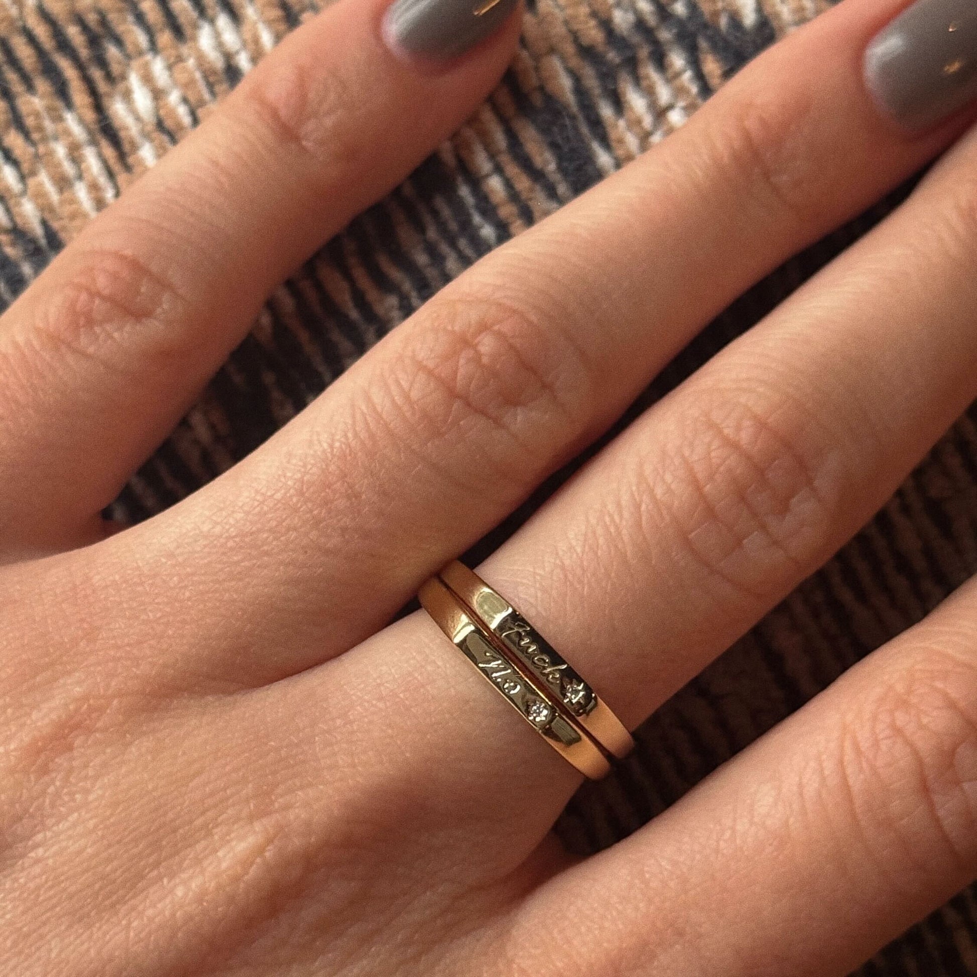 A close-up of a hand with taupe nail polish displays two 14k yellow gold plated Tiny Conversation Stacking Rings, each set with small diamonds, worn on the ring finger against a textured beige and brown background.