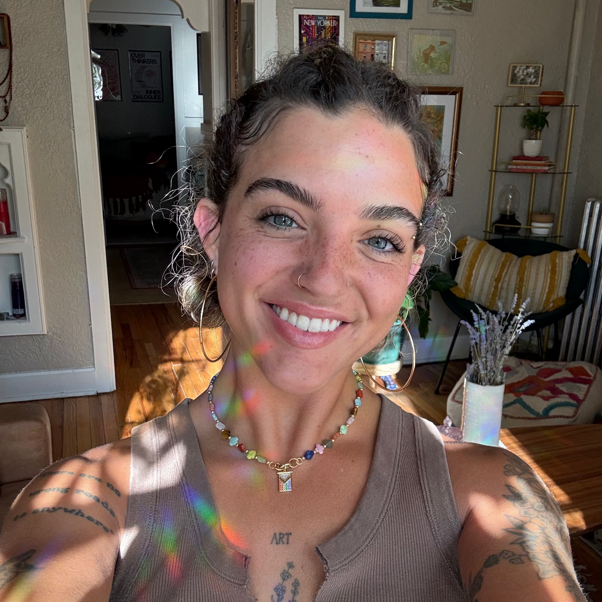 Wearing hoop earrings and a beaded necklace featuring a Limited Edition PRIDE Month Charm, a person with curly hair smiles indoors as sunlight casts rainbow reflections across their face in a cozy, art-filled room.
