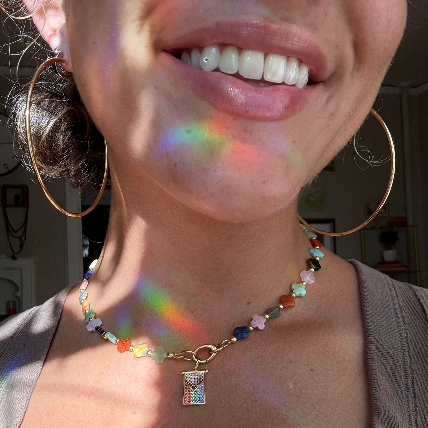 A smiling person wears large gold hoop earrings and a colorful beaded necklace featuring a Limited Edition PRIDE Month Charm, their face and neck bathed in rainbow sunlight reflections.