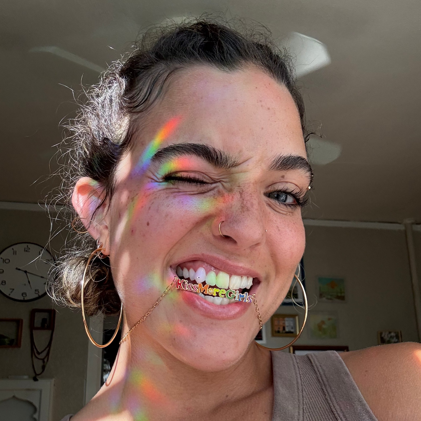 A person with large hoop earrings and a nose ring smiles and winks, holding an exclusive, colorful Kiss More Girls Necklace in their teeth. Sunlight casts rainbow streaks across their face. A clock and Local Eclectic art hang on the wall behind them.
