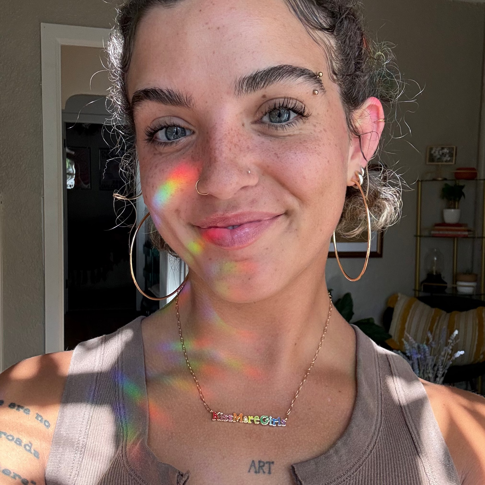 A woman with curly hair, hoop earrings, and a nose ring smiles at the camera as sunlight creates rainbow reflections on her face. She wears a Kiss More Girls Necklace from Local Eclectic, a sleeveless top, with indoor plants in the background.